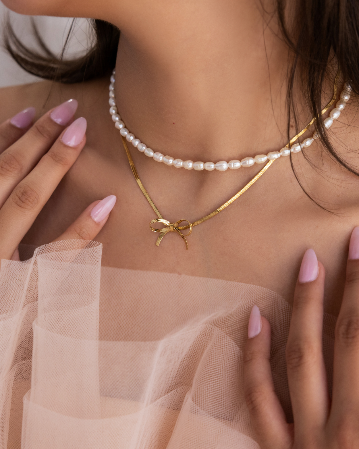 A coquette themed jewelry collection feature photo of a jewelry model wearing a beaded pearl necklace layered with a gold herringbone bow necklace.